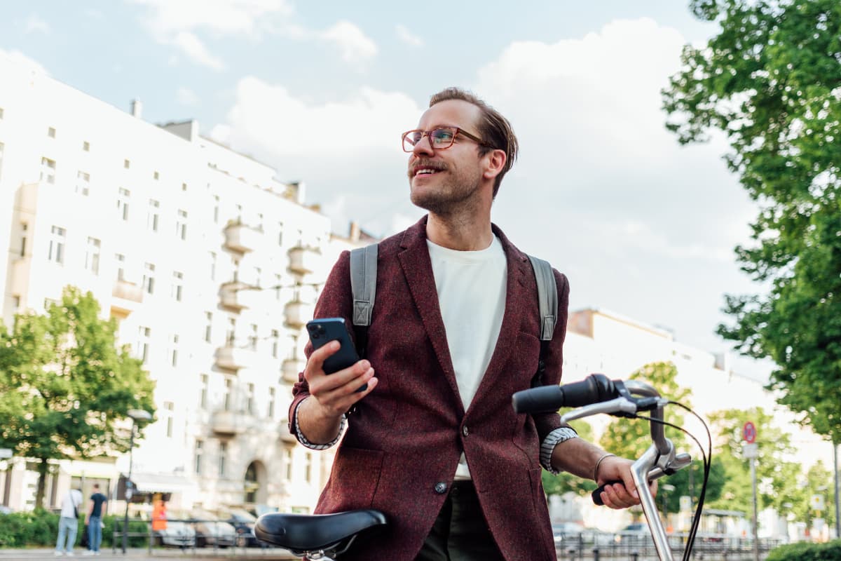 Un homme souriant tenant son vélo et son smartphone dans une rue urbaine, symbole de mobilité et de digitalisation au quotidien