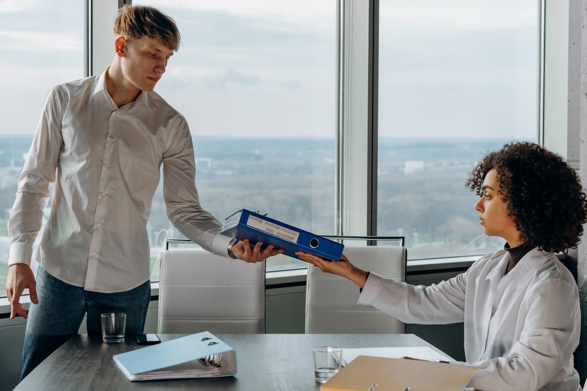 Young Man Standing in White Long Sleeve Shirt Giving a Book Report to His Coworker Inside a Conference Room